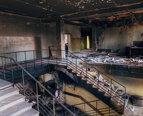 Interior of a large commercial building showing severe fire damage with charred ceilings, blackened walls, and debris across a staircase and mezzanine