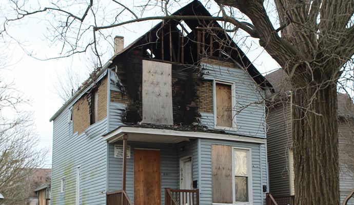 Residential property with significant fire damage and exposed roof secured with plywood board-up over windows and doors