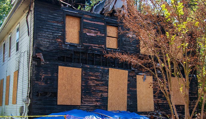 A two-story residential house with severe exterior fire damage and charred wood siding, featuring windows and doors secured with plywood boards