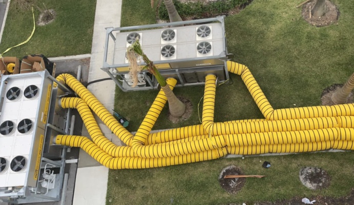 An aerial view of large industrial dehumidifiers and a network of yellow ducts set up on the grass for building restoration