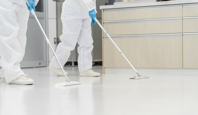 Two specialists in white protective gear mopping a white clinical floor in a kitchen or laboratory setting
